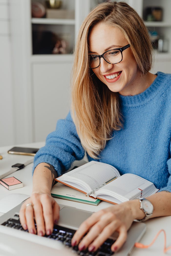 Smiling woman in eyeglasses typing on laptop at desk with open book.